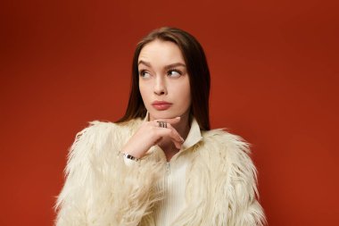 A stylish woman poses thoughtfully in a chic faux fur jacket against a bold red background. clipart