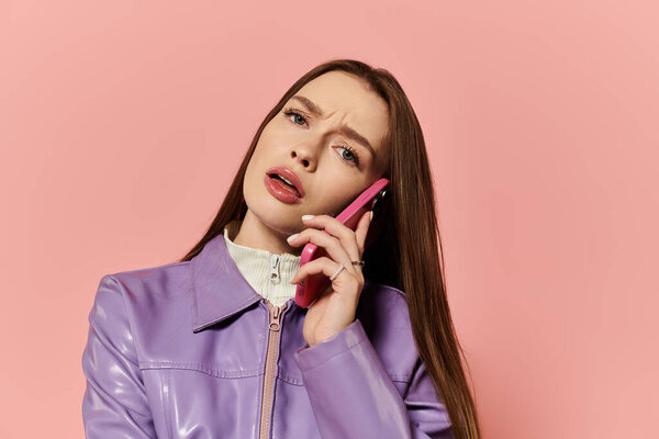 Vibrant woman in stylish outfit talks on phone against a soft pink background, showing curiosity.