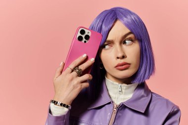 Mor kısa saçlı şık bir kadın pembe arka planda akıllı telefonuyla poz veriyor..