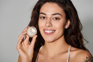 Beautiful young woman holds skincare cream, smiling confidently against a light backdrop. clipart