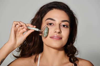 Woman enjoys skincare by using a jade roller on her face, enhancing her beauty routine at home. clipart