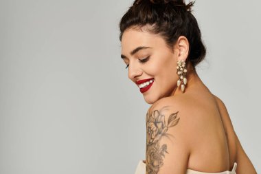 Beautiful young woman with a tattoo and striking earrings smiling confidently in a studio setting. clipart