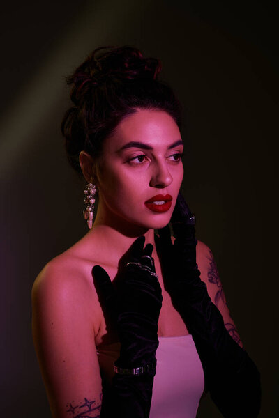 Молодая женщина излучает элегантность со смелой помадой и стильными перчатками при тусклом освещении.