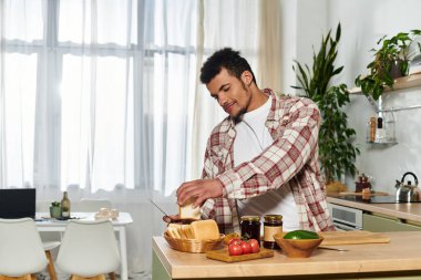 Genç bir adam ekmek dilimliyor ve bitkilerle dolu güneşli bir mutfakta sandviç süslüyor..
