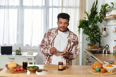 Handsome young man spreads chocolate on toast in a well lit kitchen filled with greenery. clipart