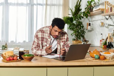 A young handsome man is working intently on his laptop while taking notes in a cozy kitchen. clipart