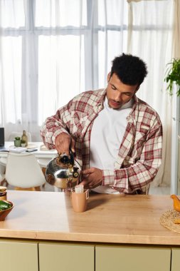 Genç yakışıklı bir adam güneşli bir mutfakta çay hazırlıyor, günlük kıyafetlerini giyiyor..