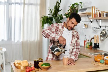 Genç adam güneşli bir mutfakta kahvaltı hazırlarken fincana kahve koyar..