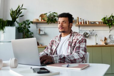 Handsome young man focuses on his laptop while seated at a stylish home office desk. clipart