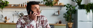 Handsome young man engages in a phone conversation while sitting in a bright kitchen. clipart