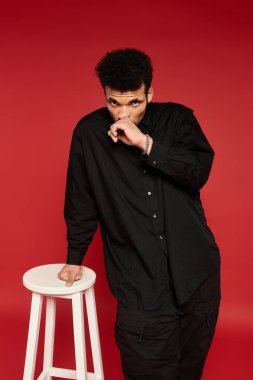 Genç bir adam beyaz bir taburede duruyor, şık bir kıyafet ve düşünceli bir ifade sergiliyor..