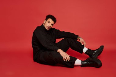 Şık bir genç adam yerde oturuyor, dikkat çekici bir kırmızı arka planla kendine güveni yansıtıyor..