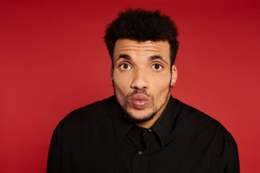 Kıvırcık saçlı genç adam şaşkın bir ifadeyle ve kırmızı arka planda poz veriyor..