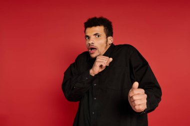 Young handsome man appears startled while posing against a bright red background. clipart