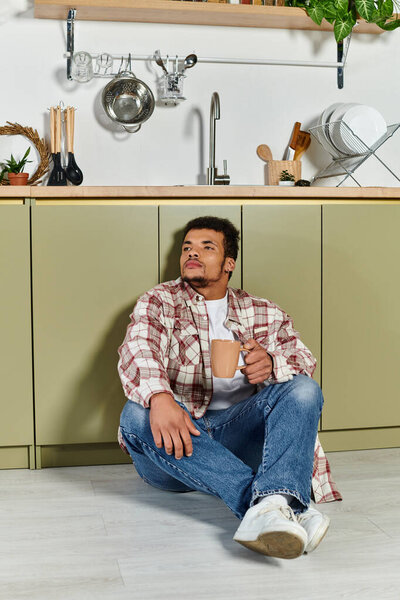 A young man relaxes on the floor of a stylish kitchen while holding a cup, looking thoughtful.
