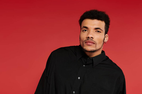 A young, handsome man stands confidently with a slight tilt against a vivid red backdrop.