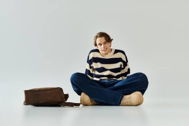 Çizgili süveter giyen genç bir adam, bir stüdyoda rahatça oturur, gençlik ve stili somutlaştırır..