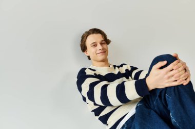 Çizgili süveter giyen genç bir adam stüdyo ortamında rahatlıyor, üniversite hayatı ve tarzı somutlaştırıyor..