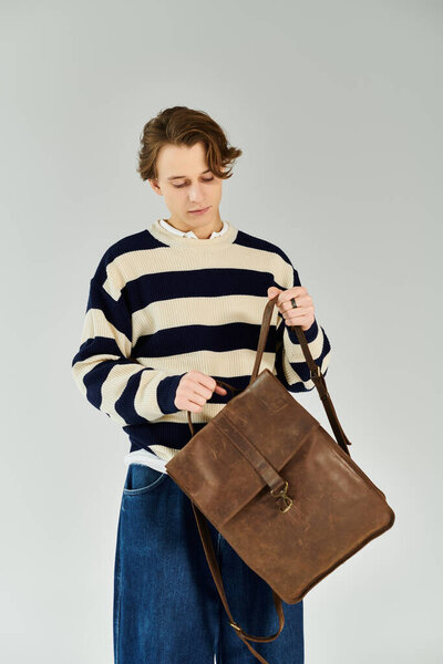 Стильный молодой человек в полосатом свитере осматривает свою кожаную сумку, готовую к поступлению в университет.