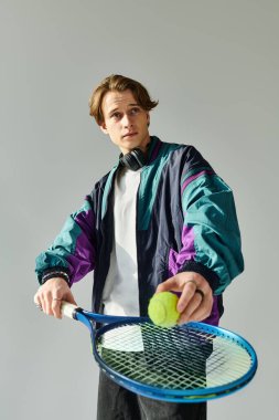 A young man in a colorful bomber jacket holds a tennis ball and racket, poised for action. clipart