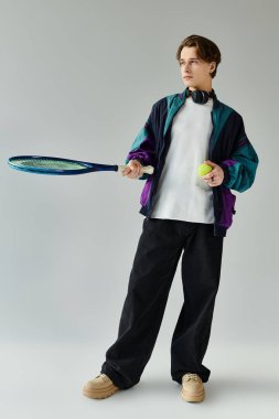 Yakışıklı genç bir adam elinde tenis raketi ve top tutarken moda tarzını sergiliyor..