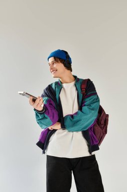 Moda giyinmiş bir üniversite öğrencisi, minimalist bir stüdyoda elinde telefonla gülümsüyor..