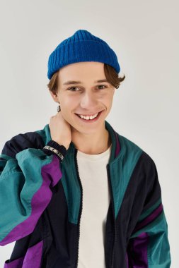 A young man wearing a colorful bomber jacket and beanie smiles brightly in a studio setting. clipart