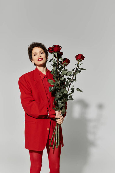 Молодая женщина уверенно стоит в ярком красном ансамбле, держа букет роз.