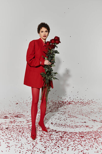 Молодая женщина уверенно стоит в ярко-красном наряде, окруженная лепестками роз.