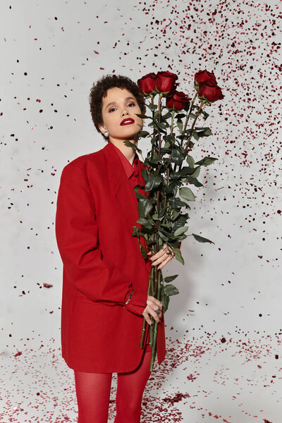Молодая женщина излучает уверенность с красными розами в руке, установленными на игривом фоне лепестков.
