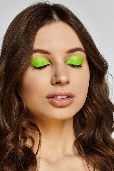 Attractive young woman poses confidently with vibrant green eyeshadow against a grey backdrop.