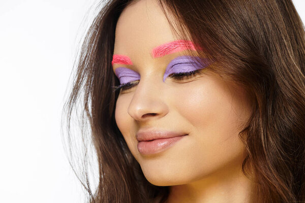 A young woman displays bold beauty with purple eyeshadow and striking pink eyebrows in a studio.