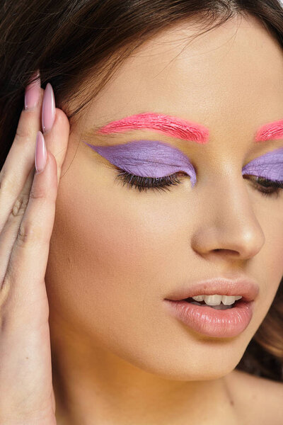 A young woman shows off bold purple eyeshadow and pink eyebrows on a grey background.