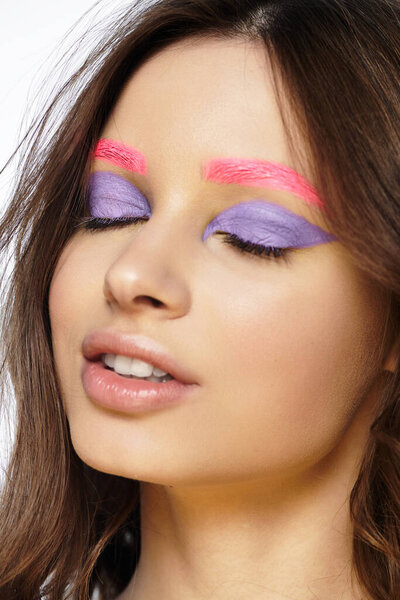 A young woman flaunts bold purple eyeshadow and pink eyebrows against a grey background.