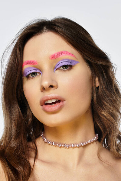 A young woman displays bold makeup with purple eyeshadow and pink eyebrows on a grey background.