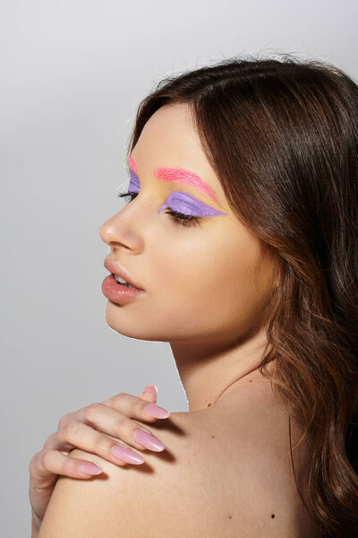 A young woman displays bold purple eyeshadow and pink brows against a grey background.