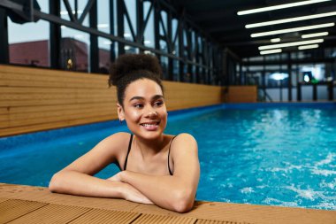 A young african american woman smiles radiantly while relaxing near a serene pool area. clipart