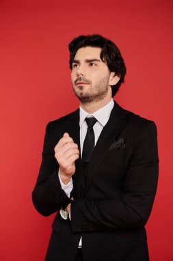 Esmer adam, siyah bir takım elbiseyle, koyu kırmızı bir duvarın önünde kendine güvenerek duruyor..