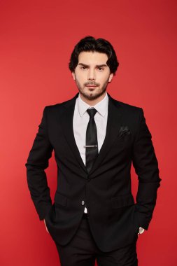 Confident young man in a black suit stands against a bold red backdrop, showcasing elegance. clipart