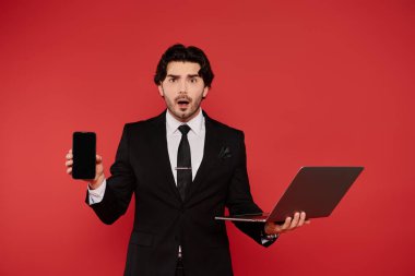 Genç adam dizüstü bilgisayarla akıllı telefonu bir arada tutarken şaşkınlık ifadesi sergiliyor..