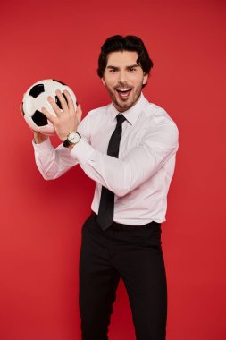 Gömlek giyip kravat takan genç ve yakışıklı bir adam futbol topuyla neşe içinde poz veriyor..