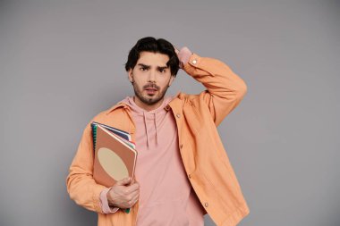 Pembe kapüşonlu ve turuncu ceketli genç bir adam elinde defterler tutarken şaşkın görünüyor..