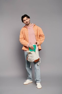 Gündelik giysiler içinde genç yakışıklı bir adam, elinde kitap ve bir sırt çantası ile güvenle ayakta duruyor..