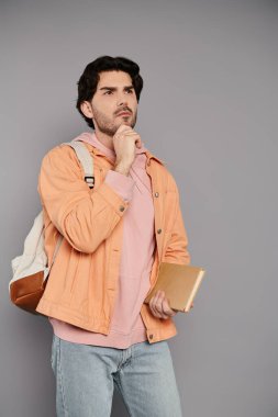 Kahverengi saçlı genç bir adam gündelik giysiler içinde elinde bir not defteri ile düşünceli bir şekilde ayakta duruyor..