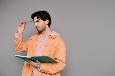 Genç bir adam, tarafsız bir arkaplan karşısında derin düşüncelere dalmış bir defter ve kalem tutuyor..