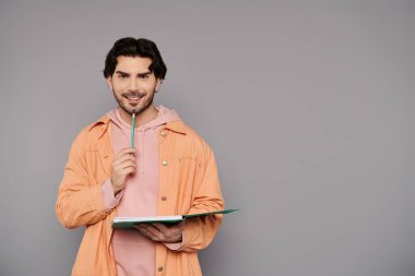 Kumral saçlı yakışıklı bir adam elinde bir defter tutarken düşünceli bir şekilde gülümsüyor..