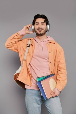 Esmer genç adam, elinde defterler ve sıradan bir atmosferde gülümserken kulaklık takıyor..