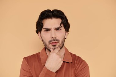 Yakışıklı bir genç adam düşünceli bir şekilde duruyor, alımlı özelliklerini yumuşak bir ışıkla vurguluyor..