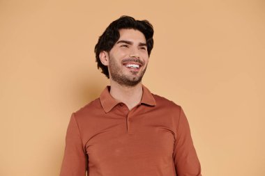 Kahverengi saçlı yakışıklı bir adam geniş bir şekilde gülümsüyor, modaya uygun bir gömlek giymiş, neşe saçıyor..