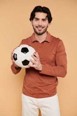 Siyah saçlı genç bir adam neşeyle gülümsüyor, elinde parlak bir ayar ile bir futbol topu tutuyor..